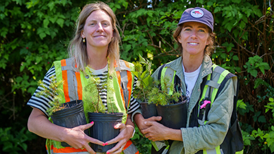 Two women in work gear holding plants with green shrubbery behind them.