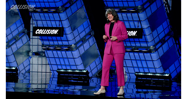 Woman in pink pantsuit on a large stage with blue lights and a sign that says Collision behind.