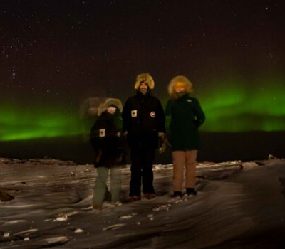 Three people in Arctic gear on the snow in front of a green Aurora Borealis.